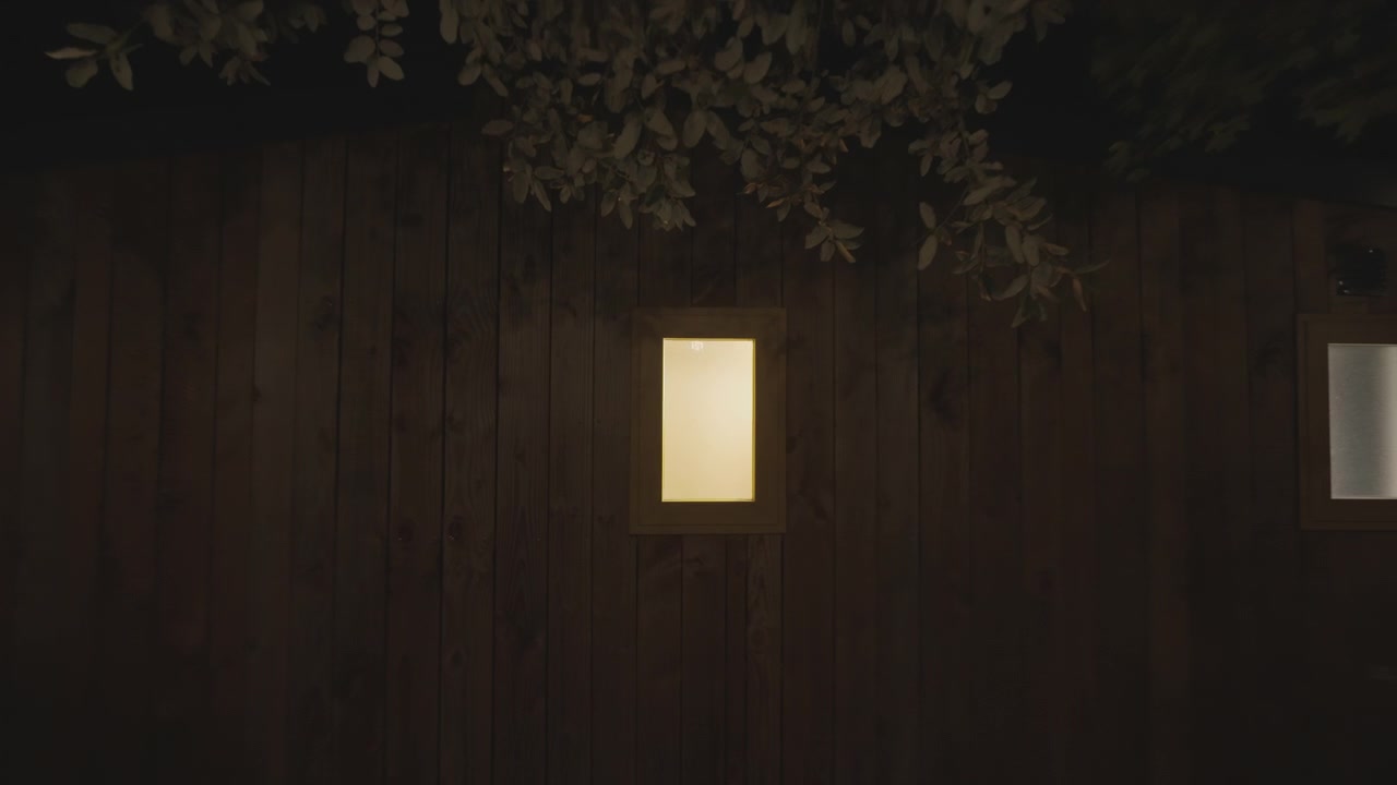 Kameraet trækker sig langsomt tilbage fra et lille, glødende rektangulært vindue på en mørk træhytte væg om natten, der afslører trægrene over hovedet