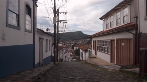 backward movement on a steep cobble stone street showing the typical colonial exterior facades of houses in ouro preto, minas gerais, brazil, with the city and mountains in the background