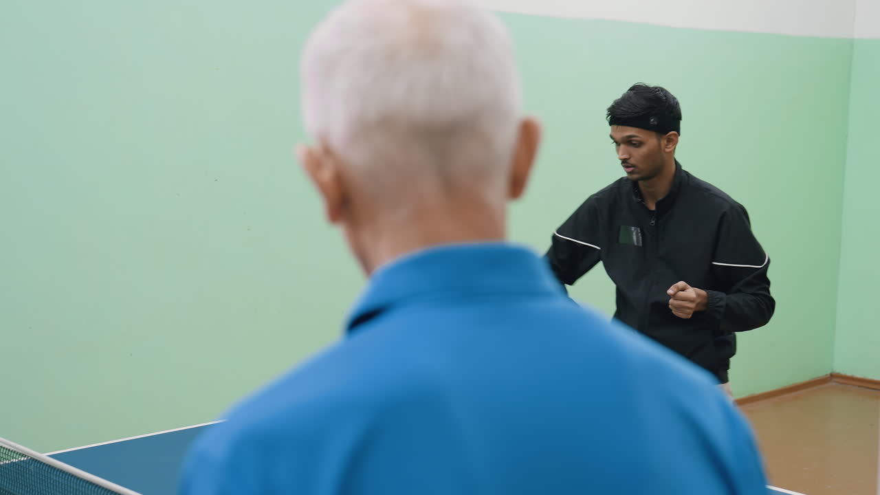 Back view blue shirt coach observes attentively as young athlete in black jacket focuses on precision and concentration while practicing table tennis with paddle during indoor sports training session