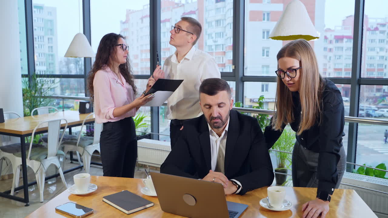 Active and busy working day in office. Boss in front of laptop and female standing next to him discussing issues in computer. Man and woman having discussion at the background.