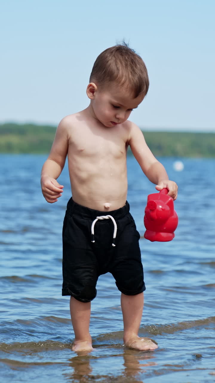 Beautiful Caucasian toddler playing in the beach. Kid carries a watering can filled with water from river. Vertical video