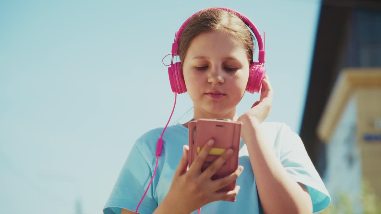 Una ragazza adolescente con cuffie rosa ascolta musica all'aperto. Tiene in mano il telefono, gli occhi fissi sullo schermo. La luce solare brillante illumina la sua espressione rilassata e felice. Perfetto per scene che riguardano gioventù e musica.