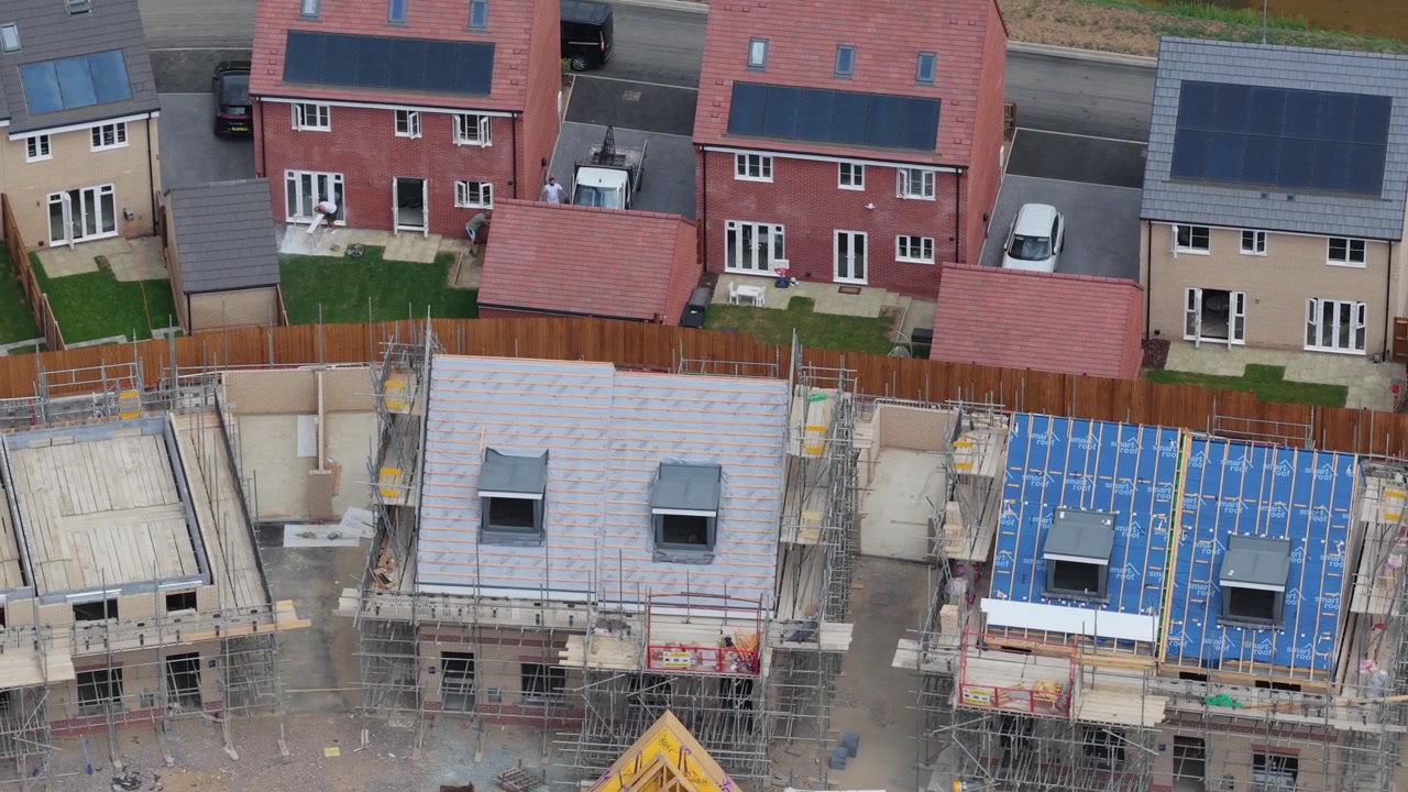 Residential area construction shows modern brick houses with roof scaffolds and blue sheeting during daylight hours as workers build new homes in a suburban neighborhood development