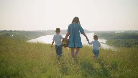 back shot of a mother in a blue dress holding a basket of fruit in one hand while gently holding her younger son's hand with the other. her older son walks beside them as they walk towards a lake