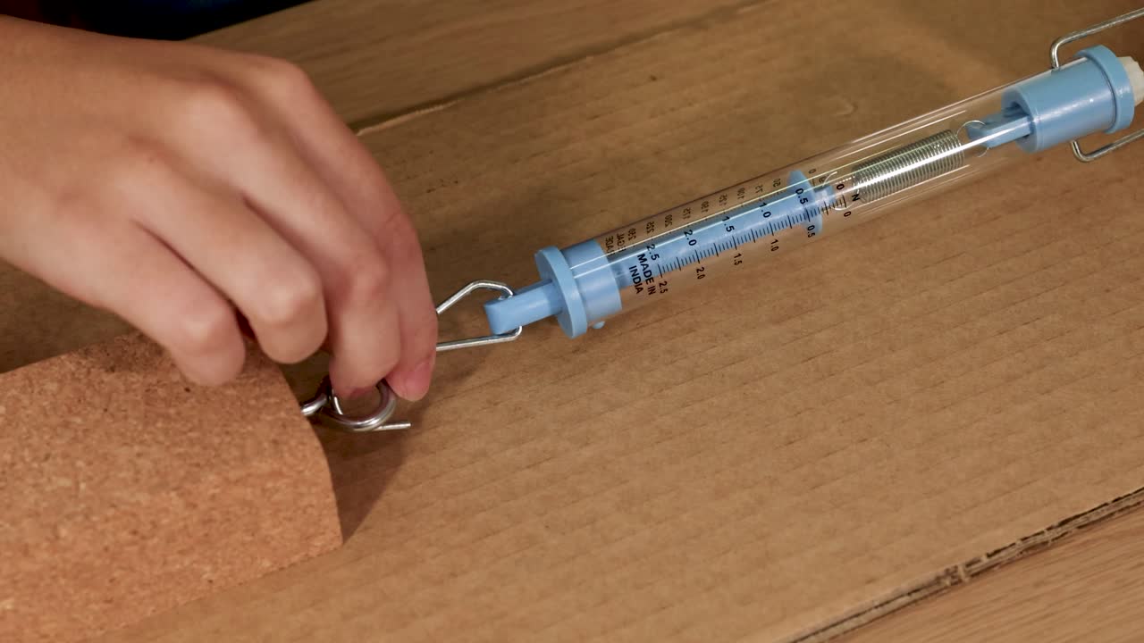 A spring scale measures frictional force as a cork block is dragged across cardboard. Hands adjust the setup