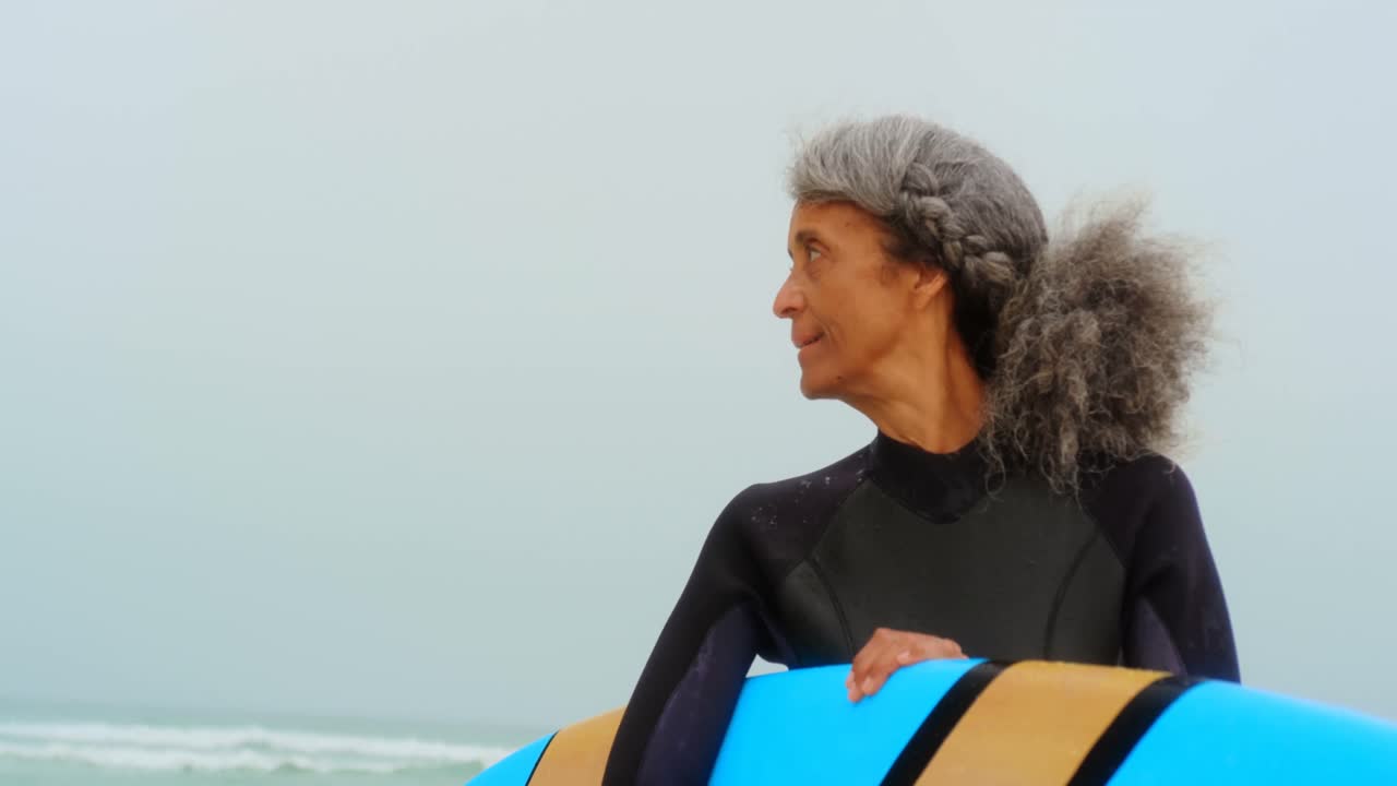 Front view of active senior African American female surfer with surfboard standing on the beach 4k