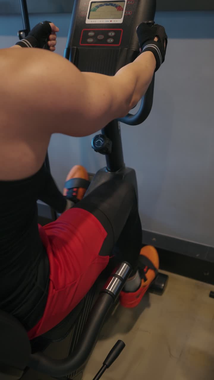 Primer plano de un hombre musculoso en pantalones cortos rojos y guantes entrenando en una bicicleta estática en un gimnasio moderno, centrado en el entrenamiento cardiovascular y el trabajo de la parte inferior del cuerpo