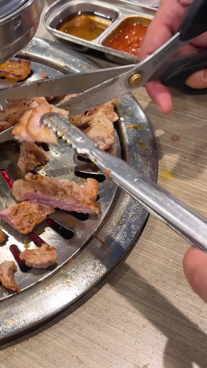 Close-up vertikal tangan menggunakan penjepit logam dan gunting untuk memotong daging babi di atas panggangan