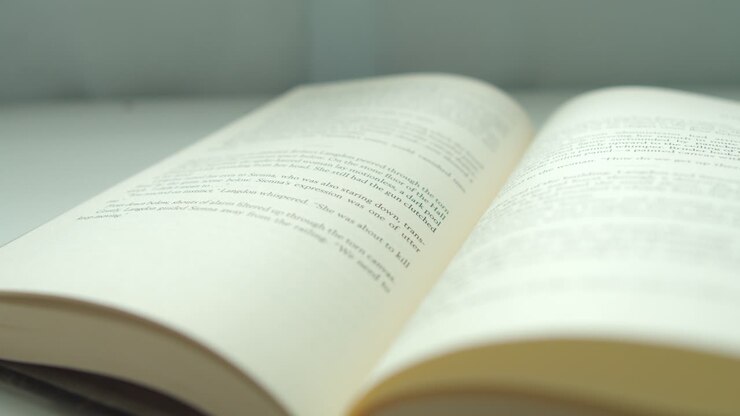 Primo piano di un dito che sfoglia una pagina di un libro da sinistra a destra, su una scrivania vicino alla finestra di casa.