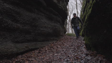 a male hiker with a red beard and a plaid flannel shirt walks roward the camers between two rocky moss covered cavernous walls