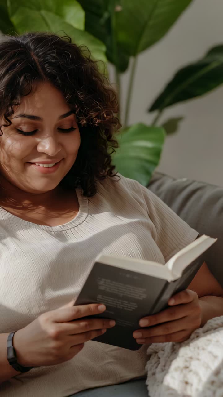 Vídeo vertical: mulher lendo um livro de bolso aberto, vestindo uma blusa clara, sentada em um sofá com uma manta e uma planta
