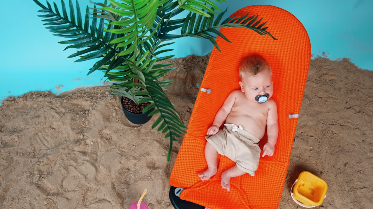 Sweet Caucasian baby boy wearing shorts lies peacefully in the chair. Beach sand and pot with plant nearby. Top view.