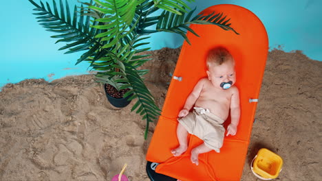 sweet caucasian baby boy wearing shorts lies peacefully in the chair. beach sand and pot with plant nearby. top view.