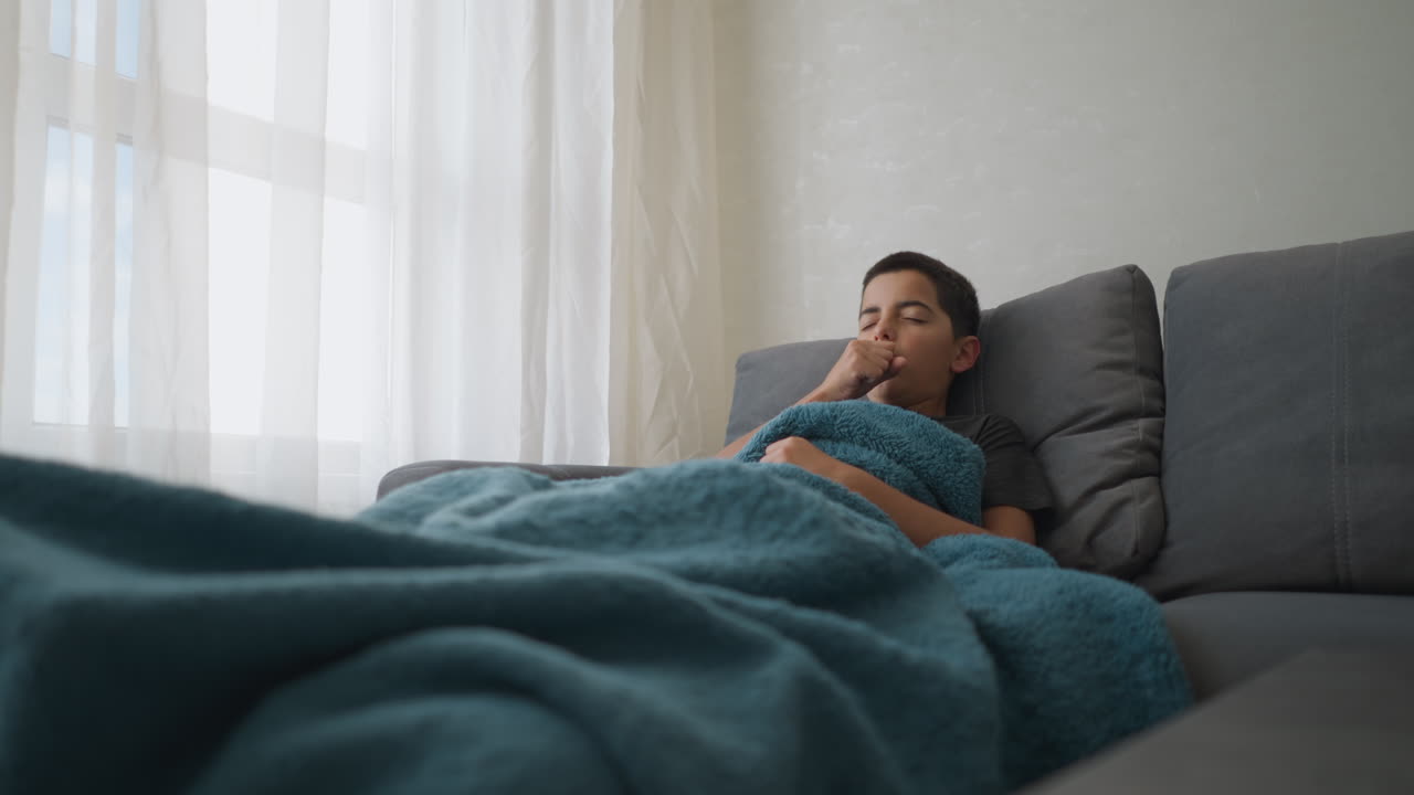 Kid lies on couch wrapped in thick blanket while coughing and covering mouth, adjusting back for comfort in softly lit modern living room
