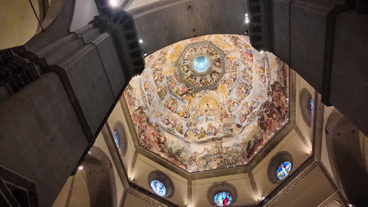 Inquadratura cinematografica dal basso all'interno di Santa Maria del Fiore, che mostra dettagli strutturali in primo piano e l'iconica cupola sullo sfondo, adornata con gli affreschi del Giudizio Universale di Vasari e Zuccari.
