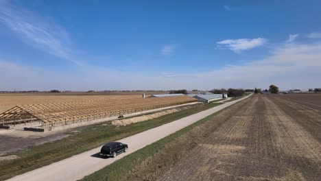 Drohnenaufnahmen-Aus-Der-Luft-Eines-Schwarzen-Geländewagens-Auf-Einer-Landstraße,-Die-An-Landwirtschaftlichen-Gebäuden-Vorbeiführt,-Unter-Dem-Blauen-Himmel-Des-Ländlichen-Iowa-Im-Mittleren-Westen