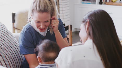 Female-healthcare-worker-with-young-mum-and-infant-son-at-home-using-stethoscope,-over-shoulder-view