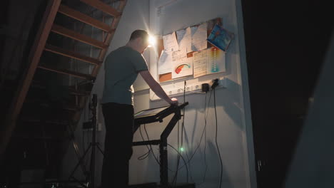 man adjusts digital control panel in dim workspace beneath staircase with wall-mounted notes and documents, standing near tripod and tangled power cords under spotlight in tech-heavy studio environment