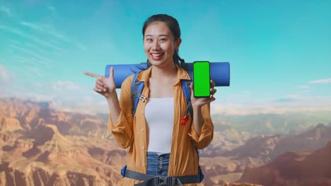 Asian Female Hiker With Mountaineering Backpack Showing Mock Up Green Screen Smartphone And Pointing To Side While Traveling At The Top Of Mountain
