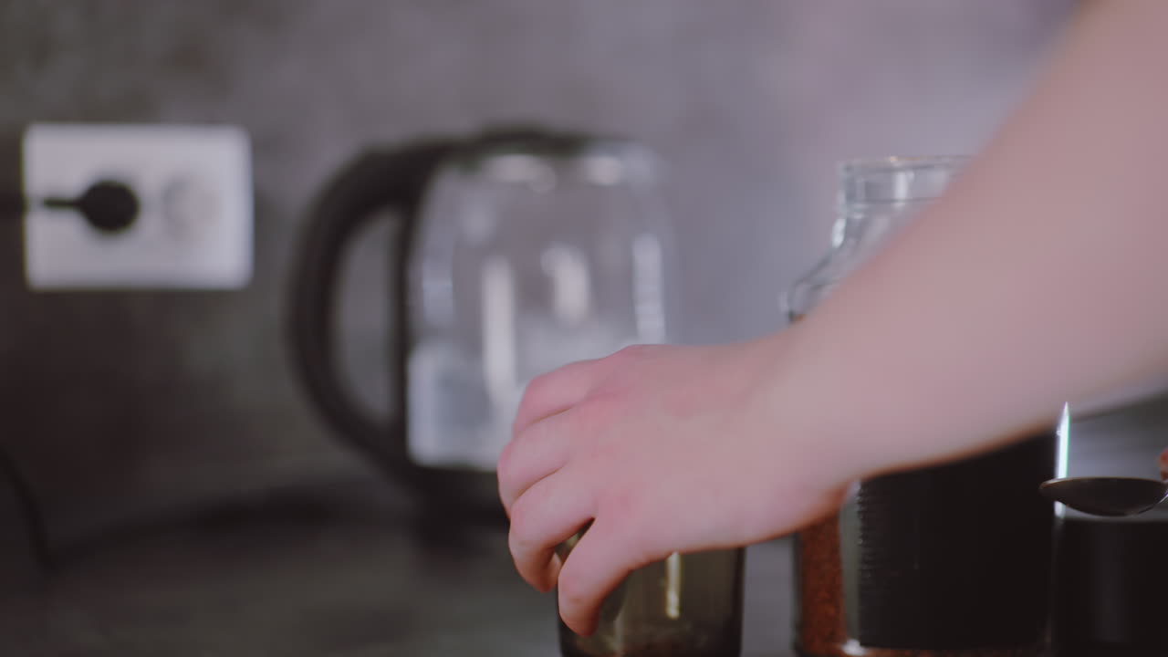 Hand pours spoonful of coffee granules into transparent glass cup while kettle boils in background, steam visible on surface, person subtly shifts cup forward on dark kitchen countertop