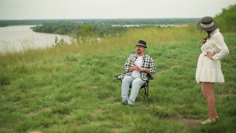 an artist in a checked shirt sits on a chair in a grassy field beside a lake, holding brushes while demonstrating a sitting pose. a woman in a white dress stands nearby, hands on hips