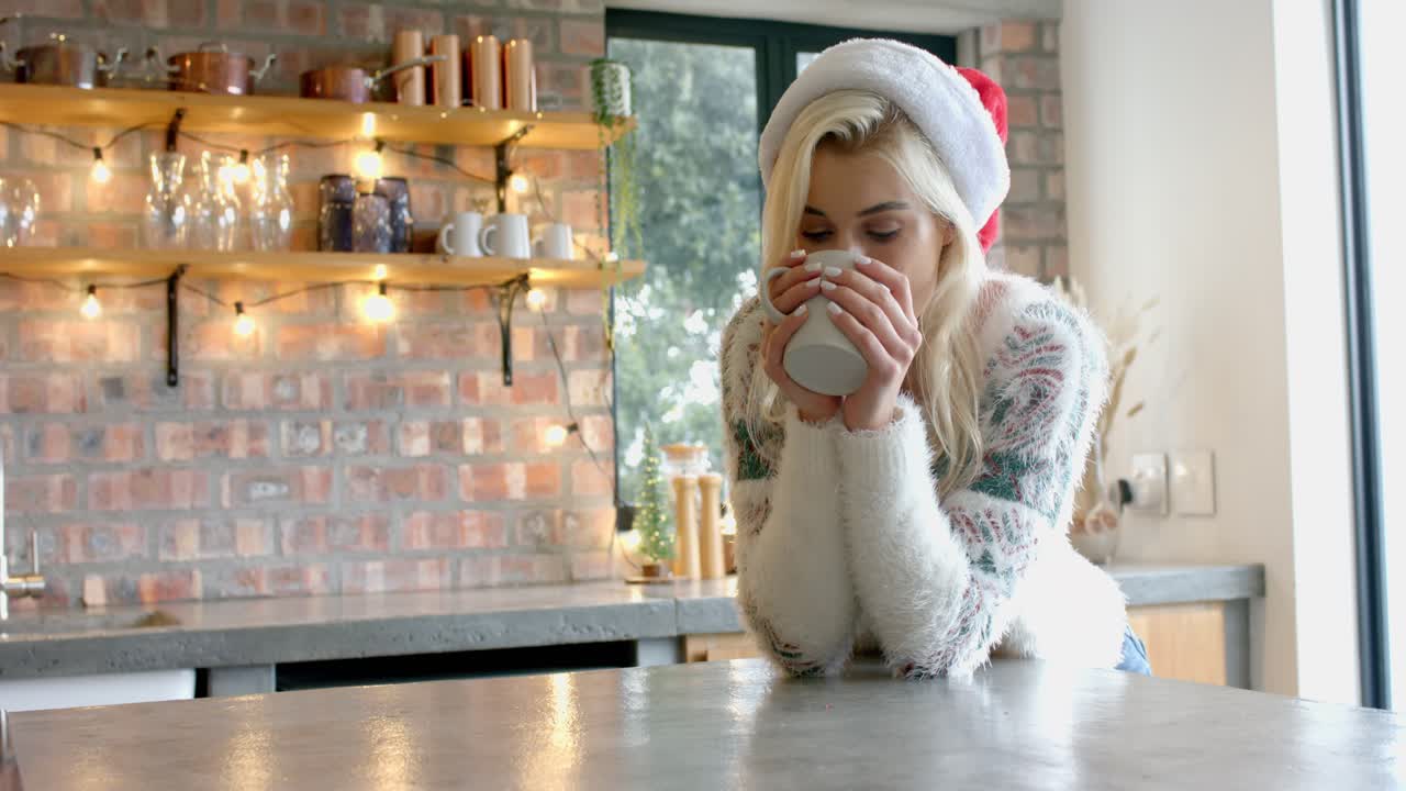 Kvinde i julesweater, der kigger ud ad vinduet, sipper for at få varmen fra et krus ved køkkenbordet, med plads til tekst.