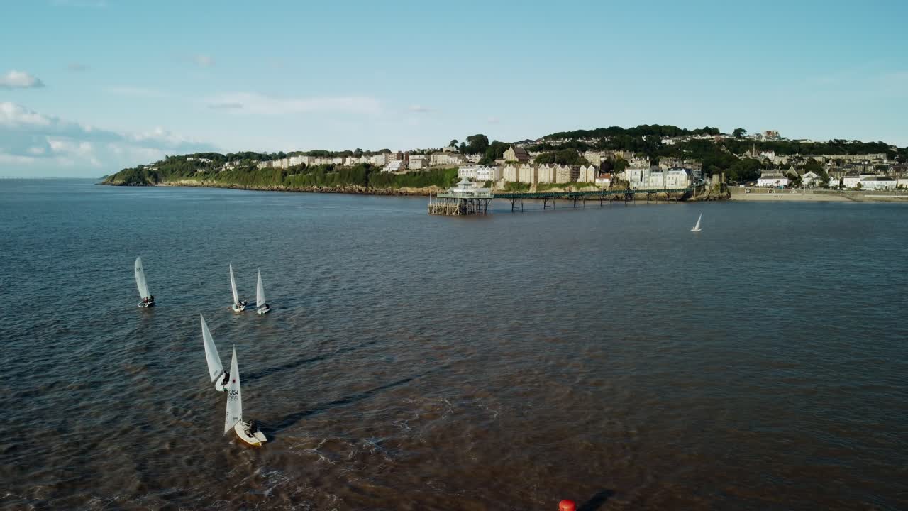 En opadgående droneoptagelse af små sejlbåde, der runder en bøje; optagelsen vipper derefter opad for at afsløre Clevedon Pier og kystlinje