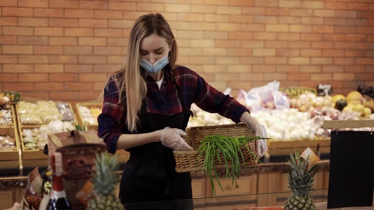 Lavoratrice con grembiule nero e mascherina che sistema le verdure nel supermercato.