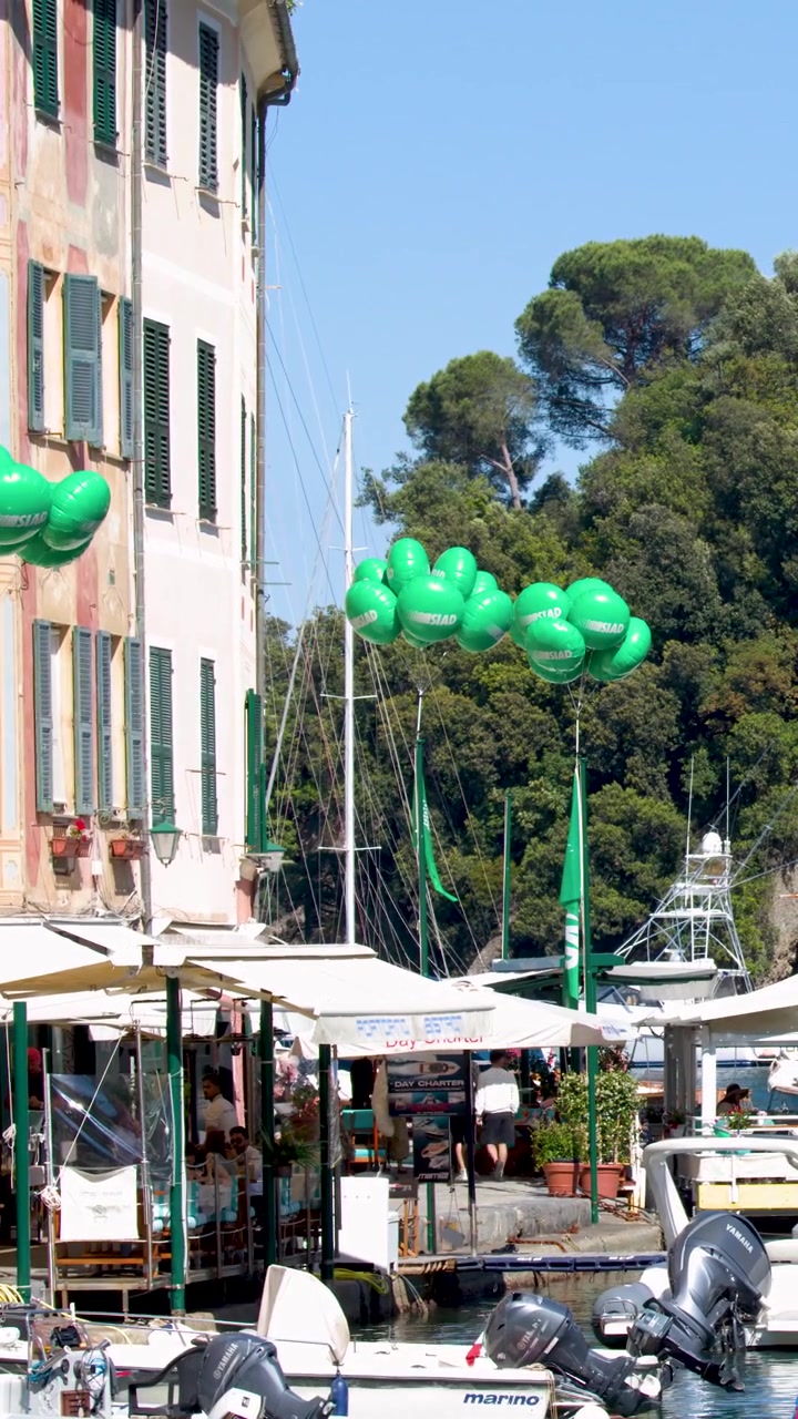 Inquadratura statica di palloncini verdi che ondeggiano sopra un vivace porto italiano con barche e caffè.