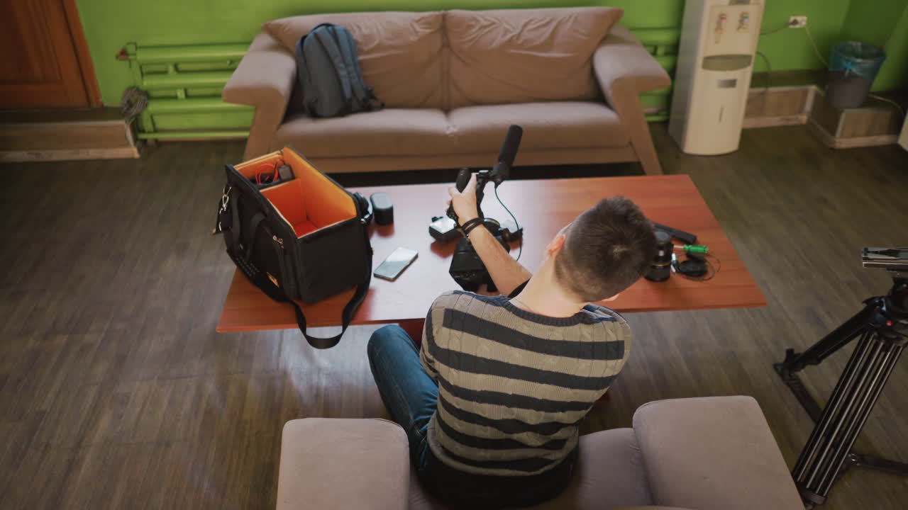Young man sets up camera equipment, removing microphone and camera gear in backpack. Casual home environment with sofa and wooden table. Professional filming preparations in cozy space