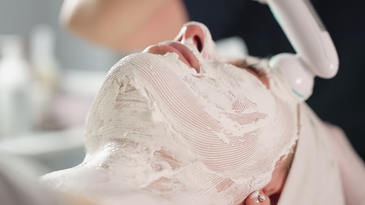 Skin therapist gloved hand maneuvers vibrating electric facial roller across white cream mask on spa client s face under bright lamp, highlighting textured mask patterns and rejuvenating treatment
