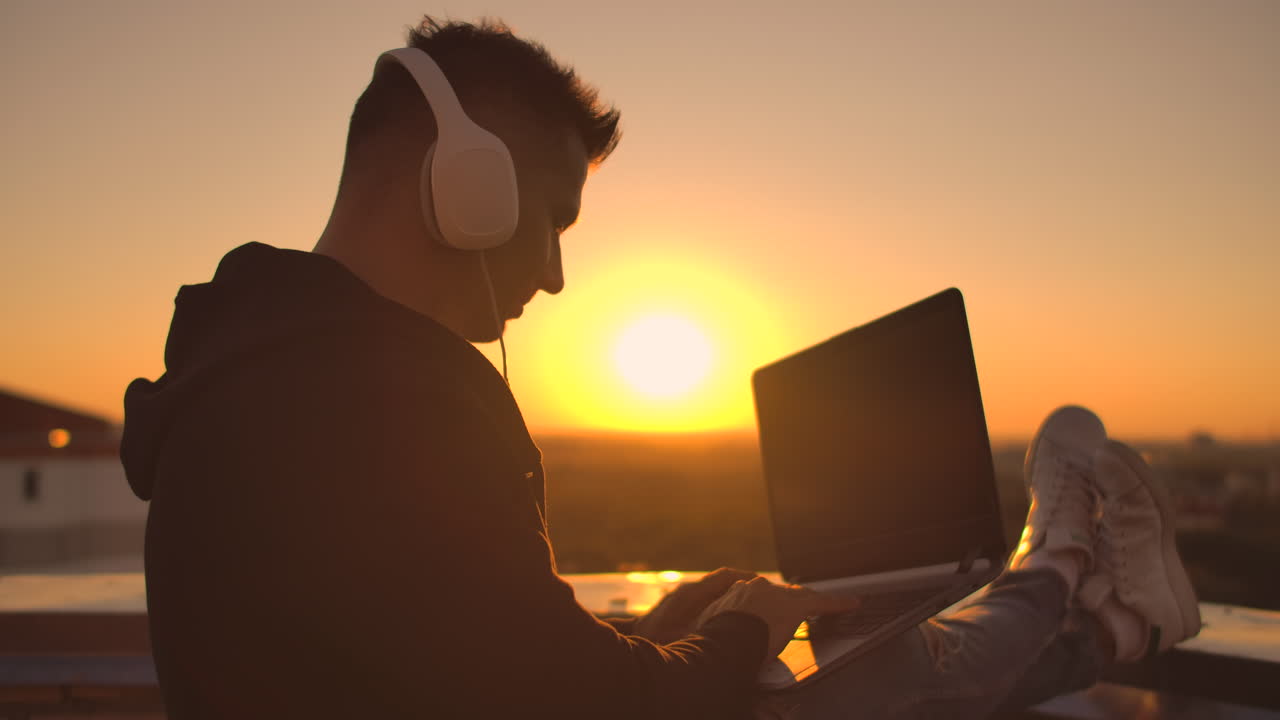 A man freelancer in headphones standing on the roof at sunset writes on the keyboard code pages. Little business. Listen to music and work at the computer enjoying the beautiful view from the roof.