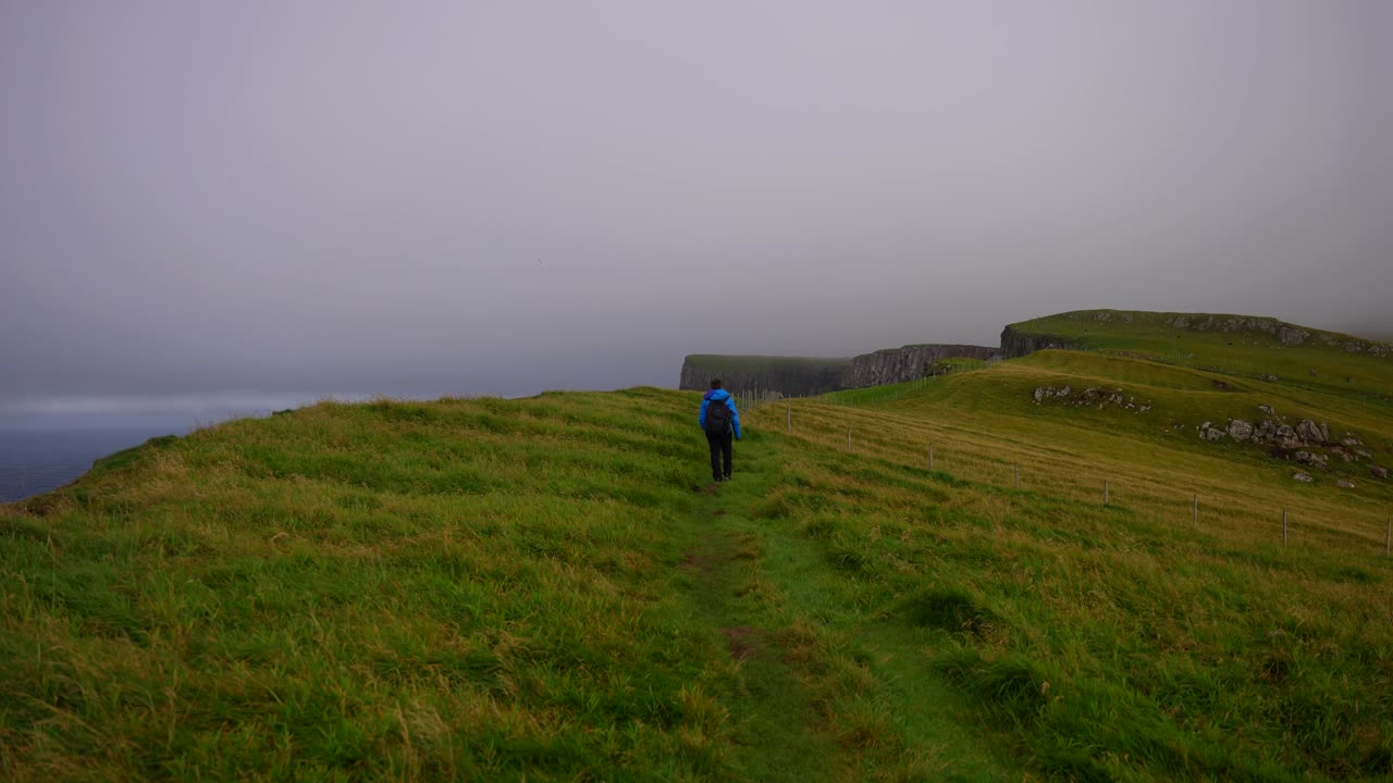 excursionista masculino camina a lo largo de la colina de hierba en mykines y se vuelve llamando a alguien en un día nublado, islas feroe