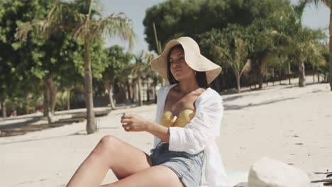Happy-hispanic-woman-in-sunhat-sitting-on-beach-in-the-sun,-copy-space,-slow-motion