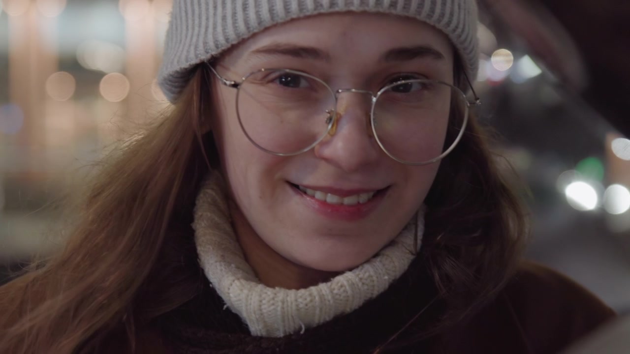 White tourist smiling at metal reflection, wearing glasses and beanie, turtleneck and coat, evening city lights, bustling plaza and railings, candid portrait shot with warm bokeh, subtle grin