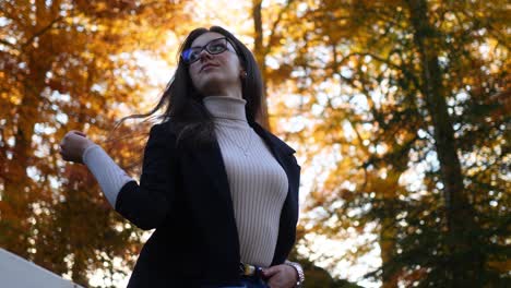 Slow-Motion-of-a-Young-Woman-on-Fence-of-Public-Park-Under-Trees-in-Fall-Colors-Touching-Her-Dark-Hair,-Low-Angle