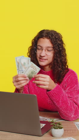 money cash dollar rain falling on girl sitting at table celebrating success on yellow background