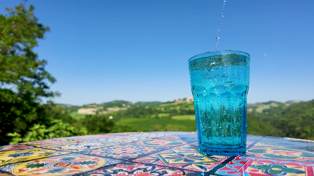 Clear sparkling water pours from a green bottle into a blue glass on sunny terrace.
