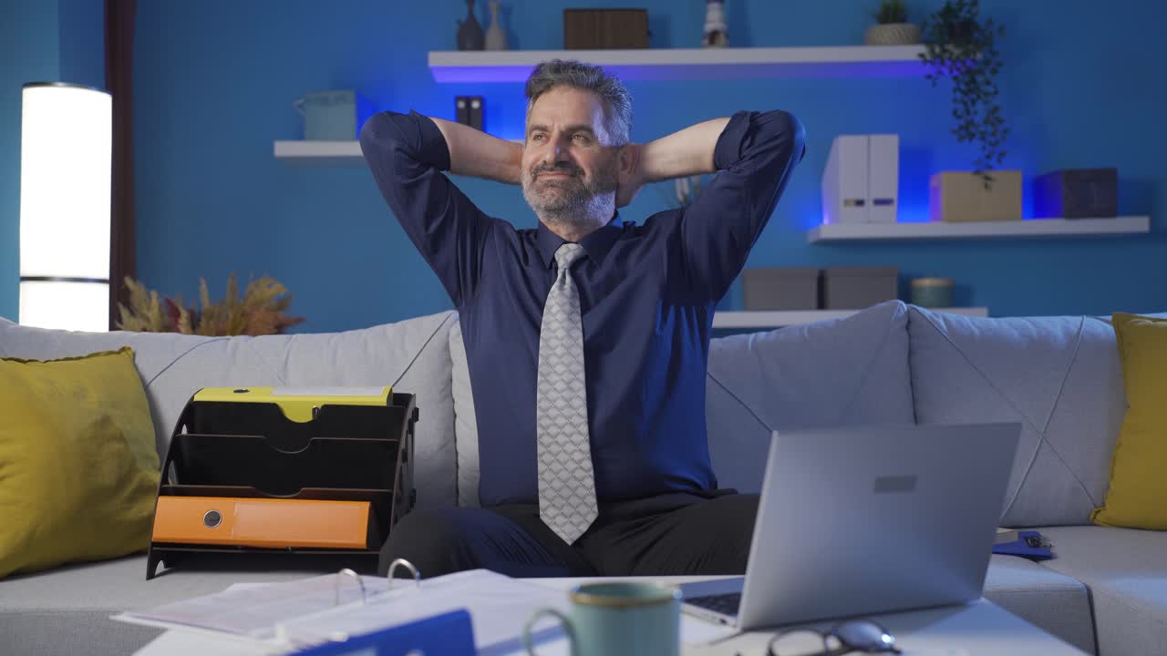Calm and successful businessman completing his online work from laptop at night at home and relaxing, being happy.