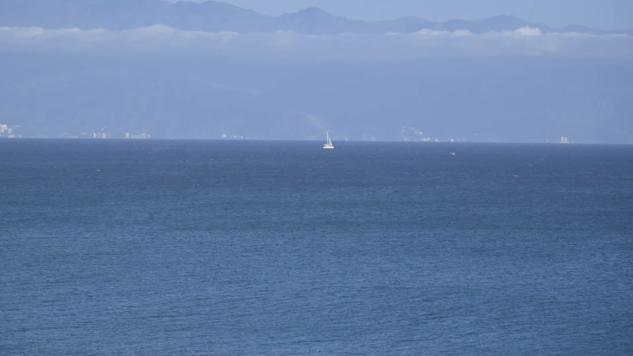 遠景に山々がそびえる外洋で、穏やかな青い海、重層的な海岸線、そして平穏な海洋の雰囲気を映し出すシネマティックなヨットの映像。