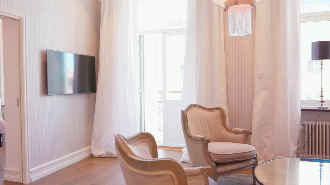 Wide gimbal shot of an elegant living room featuring vintage armchairs glass coffee table crystal chandelier floor-to-ceiling curtains bright natural light streaming windows creating airy luxurious