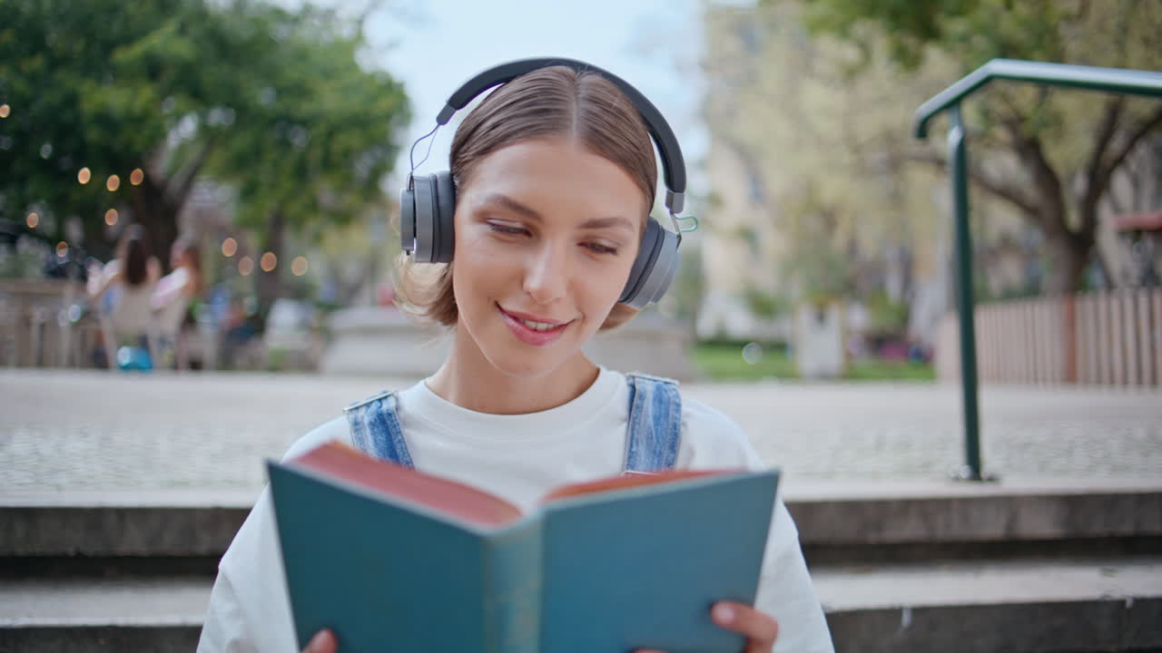 Carefree student holding book sitting street stairs in earphones closeup