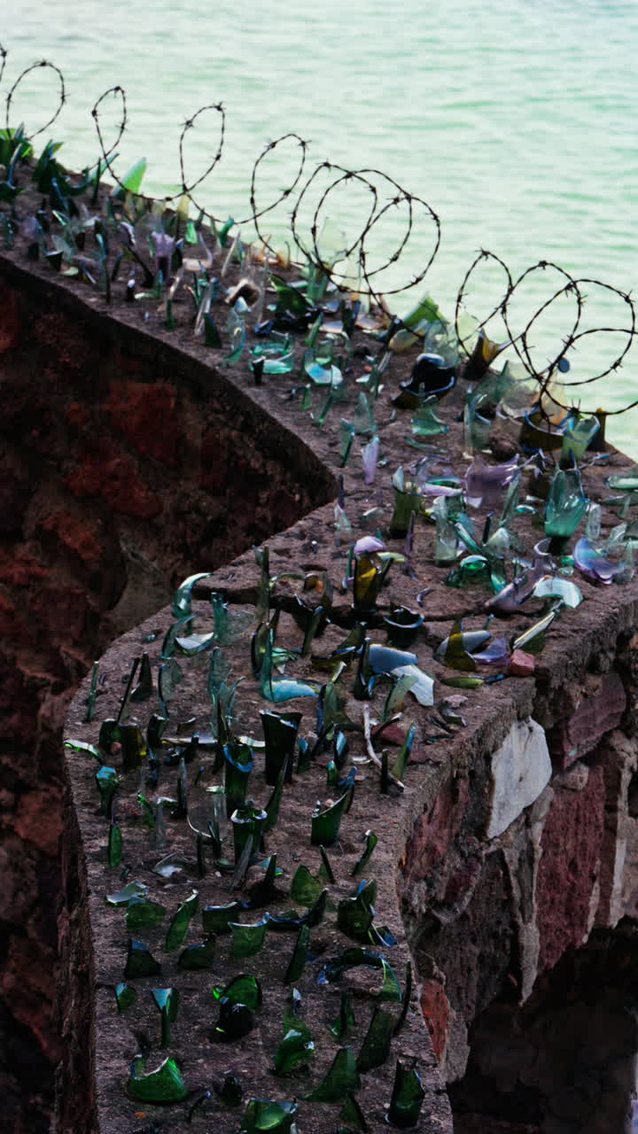 Close up of multiple broken glass shards and barbed wire loops along the edge of a brick wall with a blurred view of the sea. Vertical