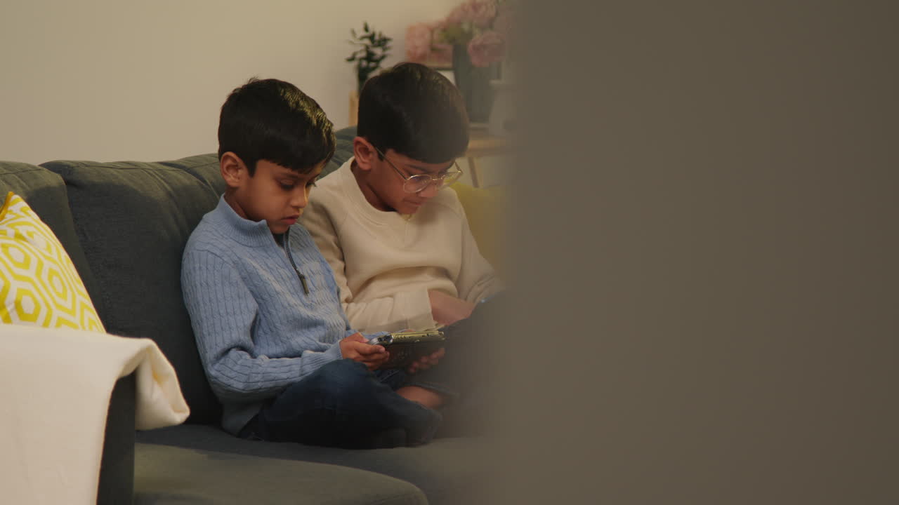 Two Young Boys Sitting On Sofa At Home Playing Games Or Streaming Onto Digital Tablets 11