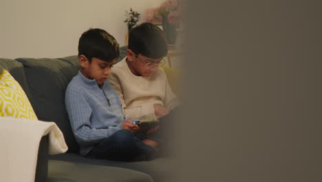 two young boys sitting on sofa at home playing games or streaming onto digital tablets 11