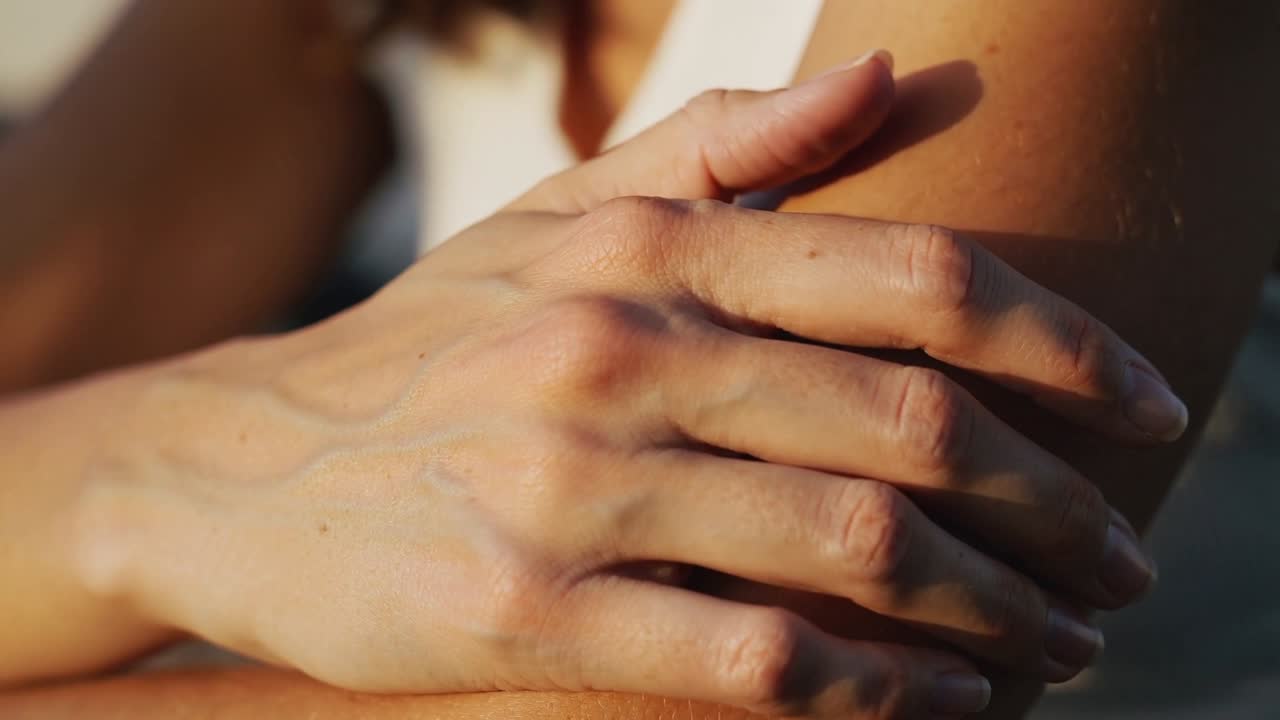 Placing hand, adjusting fingers on left forearm on patio, showing white thin strap top and veins