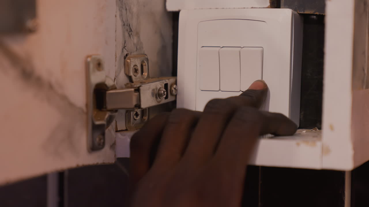 Dark hand pressing white switch closeup, fingertip engages plastic electrical switch inside aged cabinet hinge, subtle grime and rust, moody dim light, tactile domestic routine and quiet maintenance