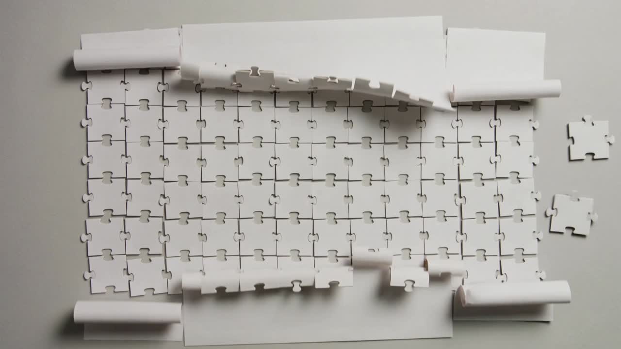 Unrolling from top, large jigsaw paper sheet revealing puzzle grid on gray table, scattering pieces