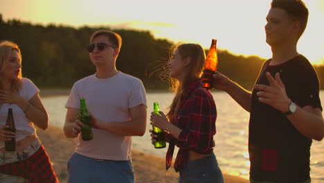 the teens spend the carefree summertime at sunset on the sand beach with beer. they are talking to each other and and laugh at funny stories on the open air party.