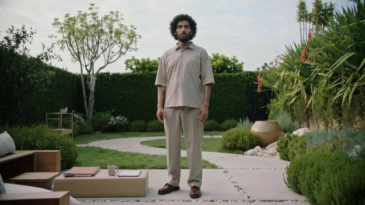 La telecamera si allontana rivelando un uomo in abito beige in piedi sul patio che mostra il giardino e una tazza
